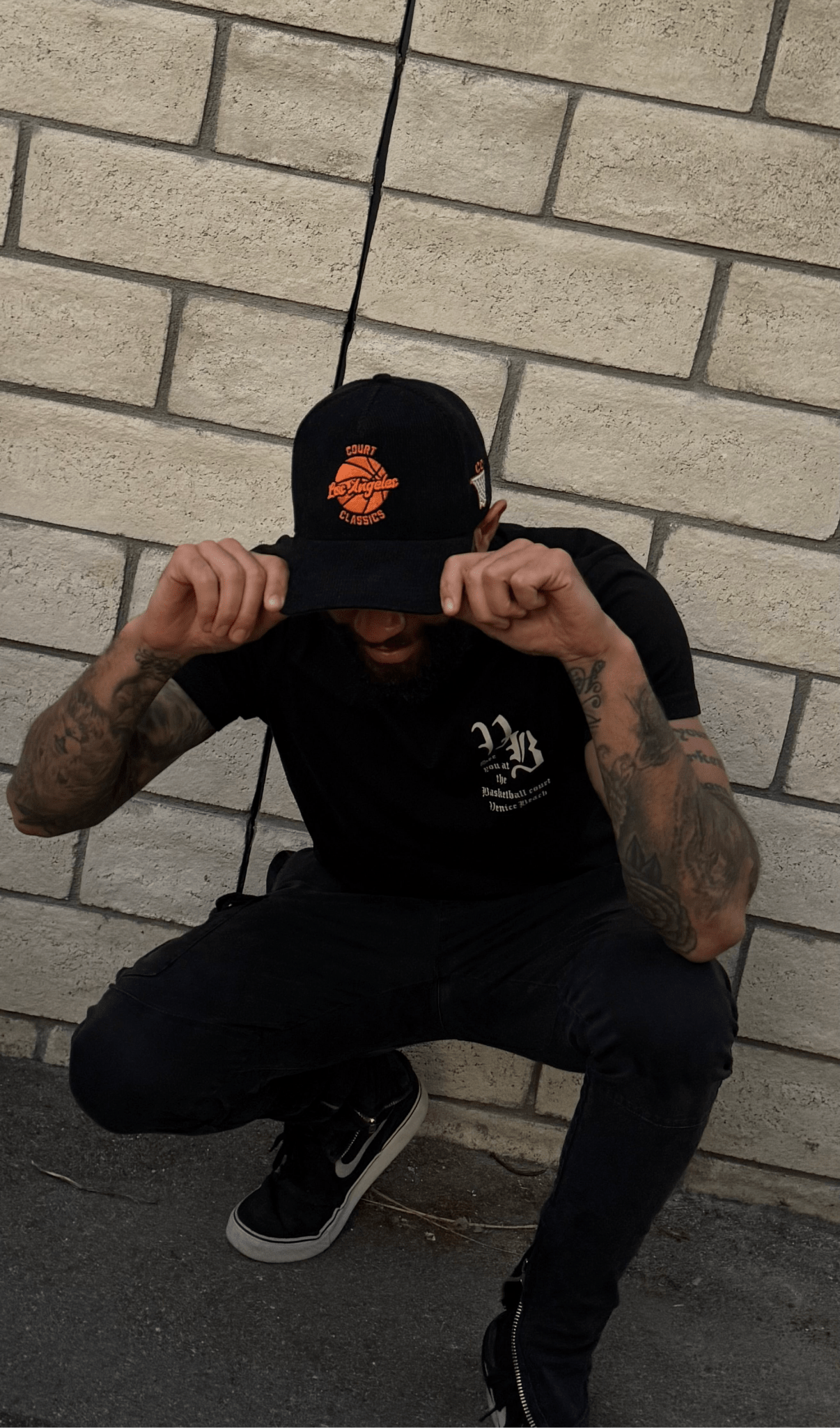 Streetwear style shot of black & orange corduroy snapback hat worn by man crouching near brick wall