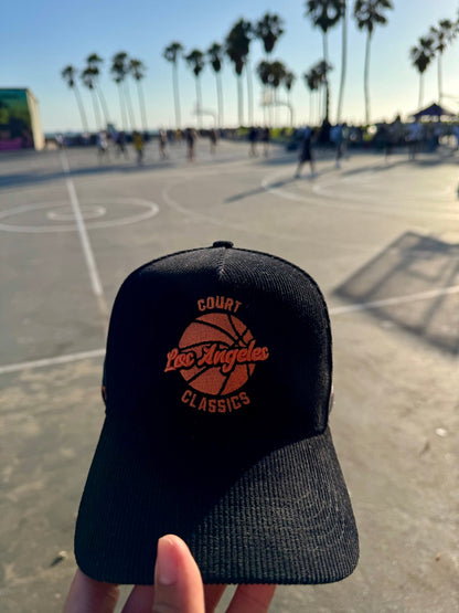 Black & orange corduroy snapback hat held in hand with basketball court and palm trees in background – Court Classics